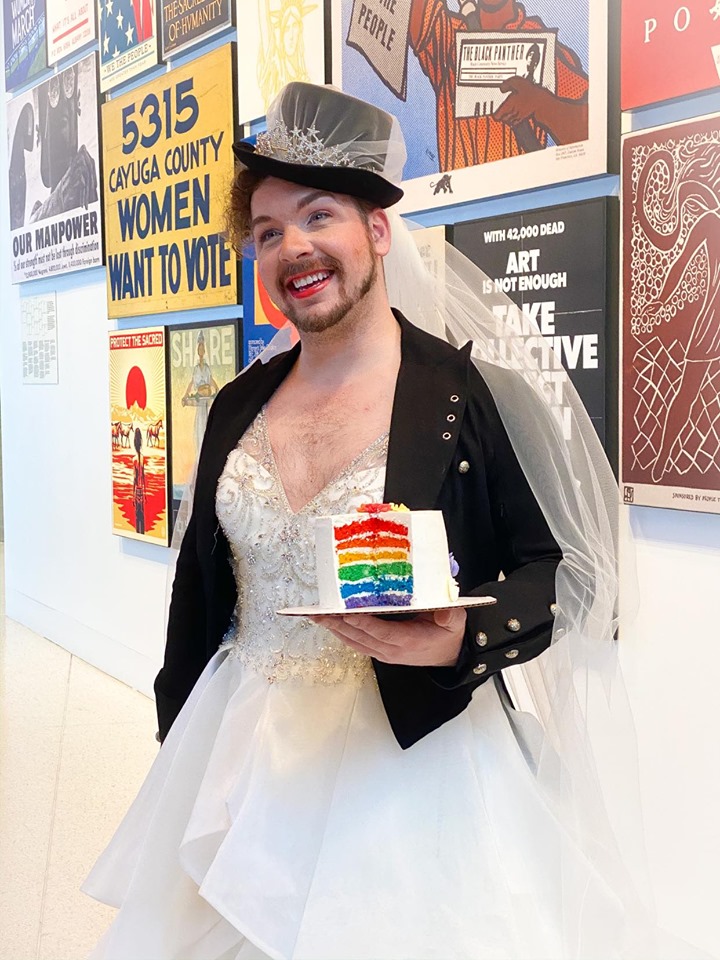 Mirror Mirror Inc. LGBT bride to be posing in their wedding gown, top hat veil and holding a rainbow wedding cake.