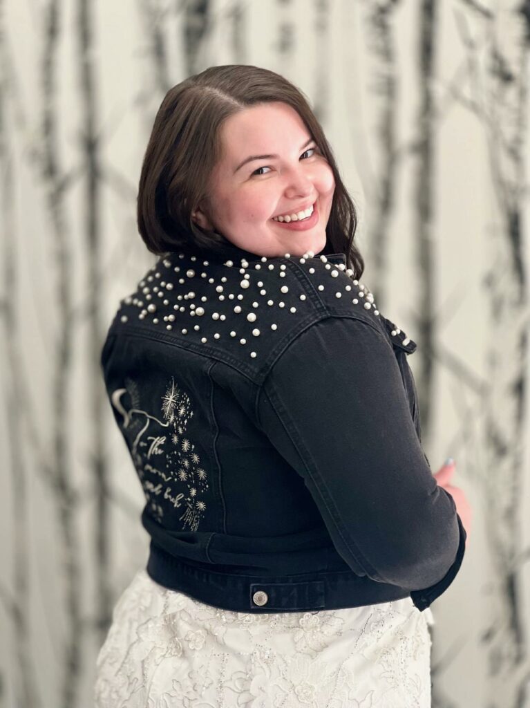 Smiling bride posing in her wedding dress and black jean jacked with pearls on the shoulders.