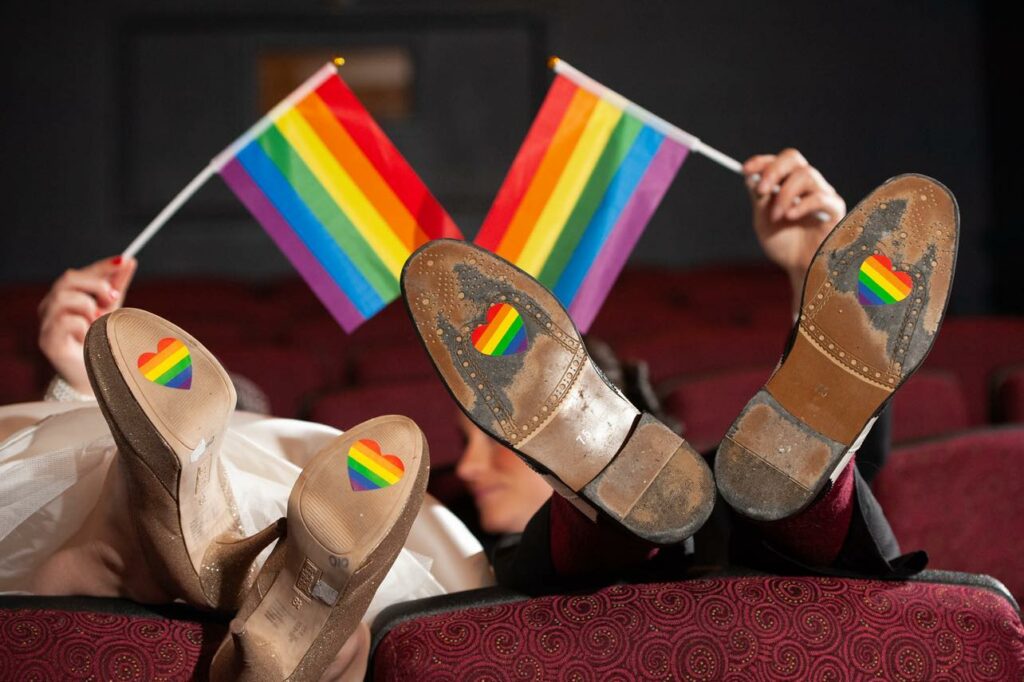 partners laying down together holding up LGBTQ flags and LGBTQ colors on bottom of shoes