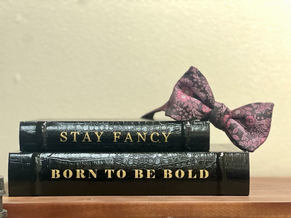 Display of books that say "Stay Fancy" and "Born to be bold" with a pink floral printed bowtie from Mirror Mirror Tux.