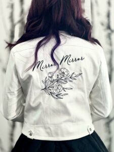 Bride to Be posing with her back to the camera to show off her white leather jacket embroidered with "Mirror Mirror" and a peony on the back.