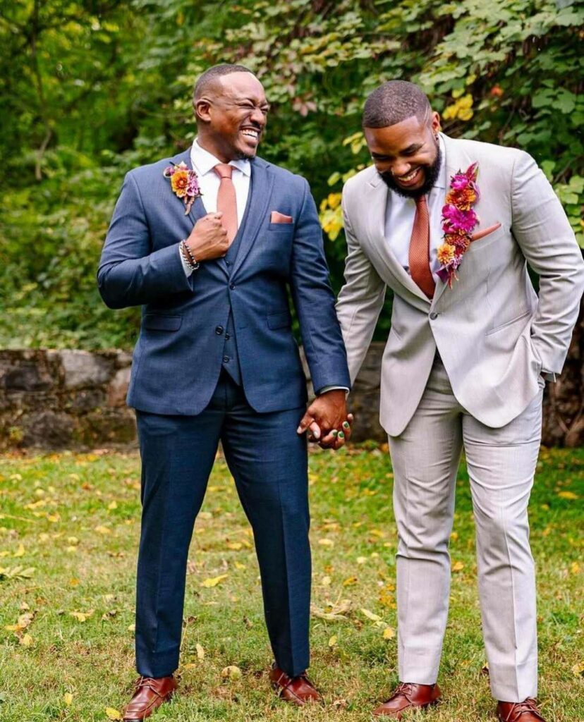 Handsome grooms standing side by side in their navy blue tuxedo and gray tuxedo wedding suits and holding hands.