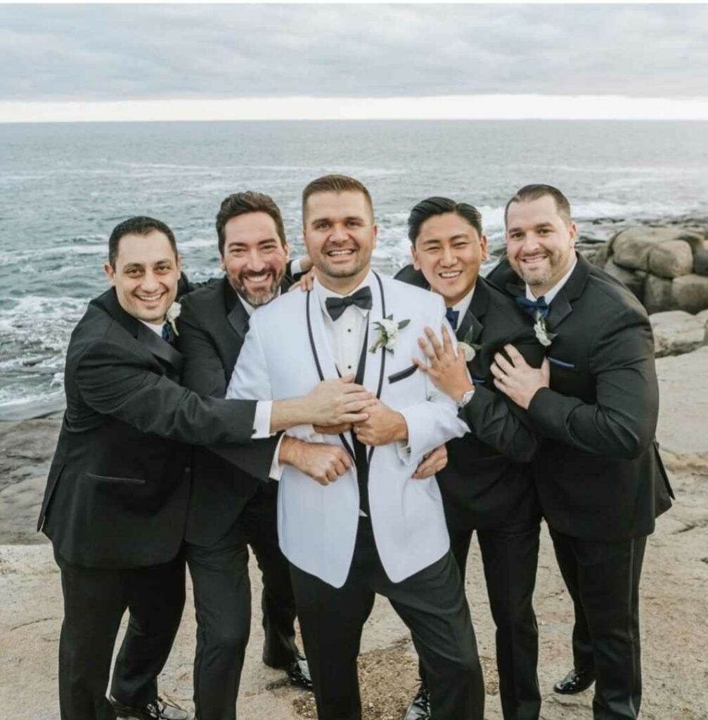 Groom and groomsmen posing together and embracing on a beach in front of the ocean.